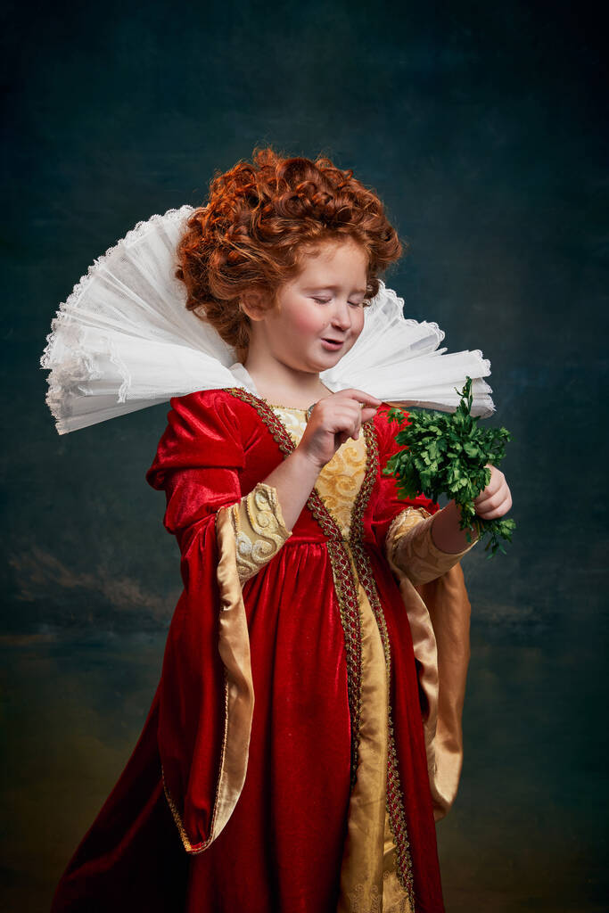 Klein roodharig meisje, kind in kostuum van koninklijke persoon met peterselie geïsoleerd op donkergroene achtergrond. Vegetariër. Concept van historische remake, vergelijking van tijdperken, middeleeuwse mode, emoties, koningin - Foto, afbeelding