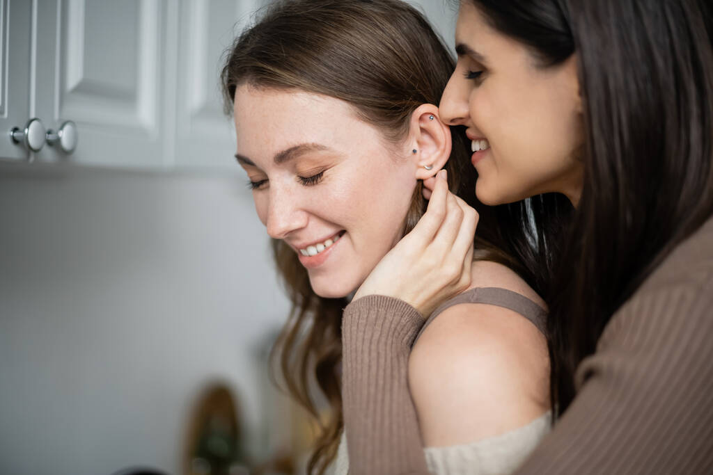 Позитивная женщина трогает ухо подруги на кухне дома  - Фото, изображение