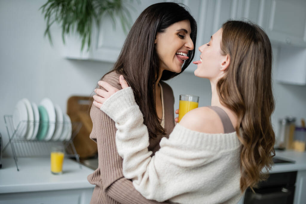 Portakal suyu içen lezbiyen kadınlar mutfakta dillerini çıkarıyorlar.  - Fotoğraf, Görsel