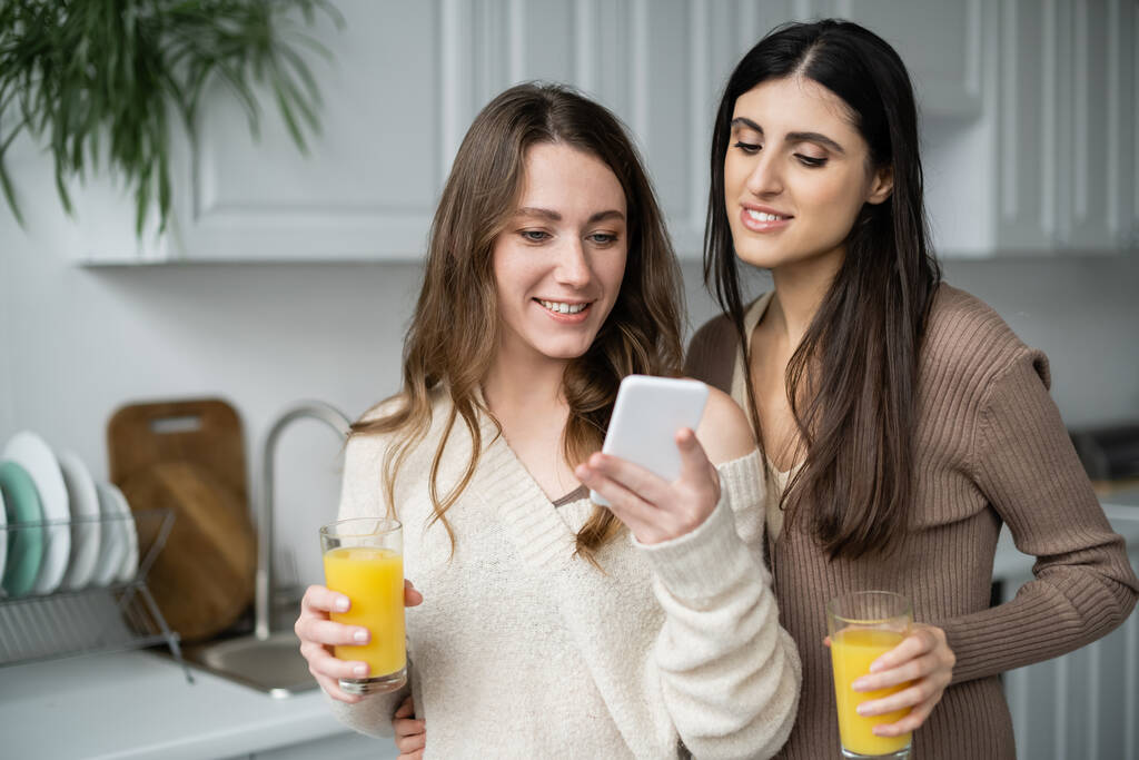 Молодая женщина держит апельсиновый сок и использовать смартфон рядом с подругой на кухне дома  - Фото, изображение