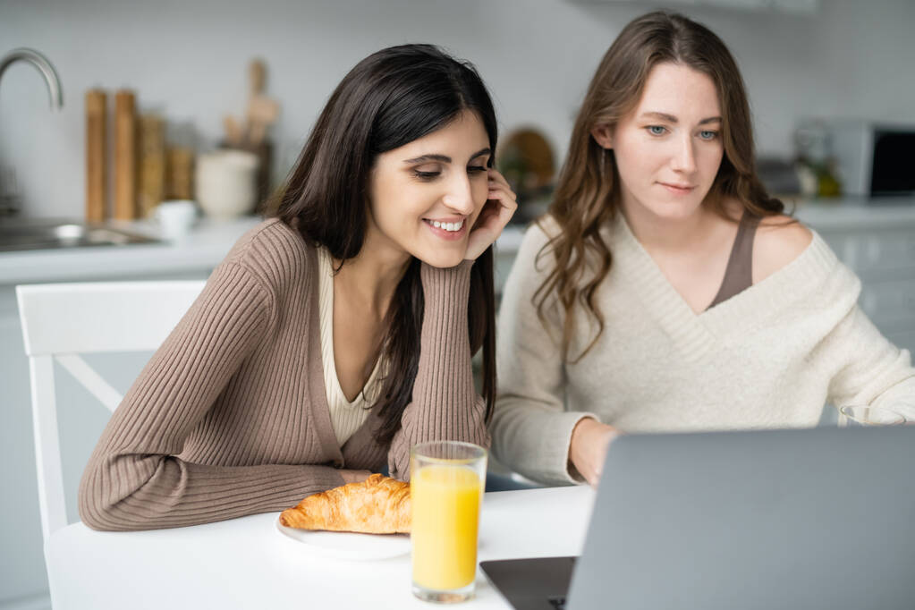Mujer lesbiana mirando borrosa portátil cerca del desayuno y novia en la cocina  - Foto, imagen