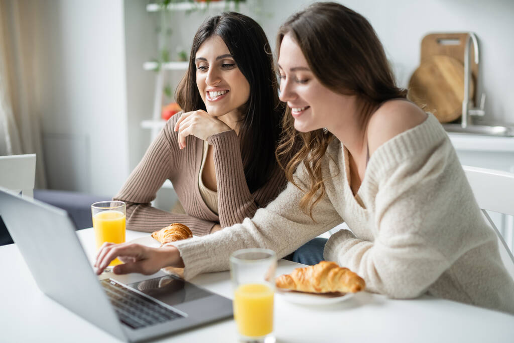 Cheerful same sex couple using laptop near croissants and orange juice in kitchen  - Photo, Image
