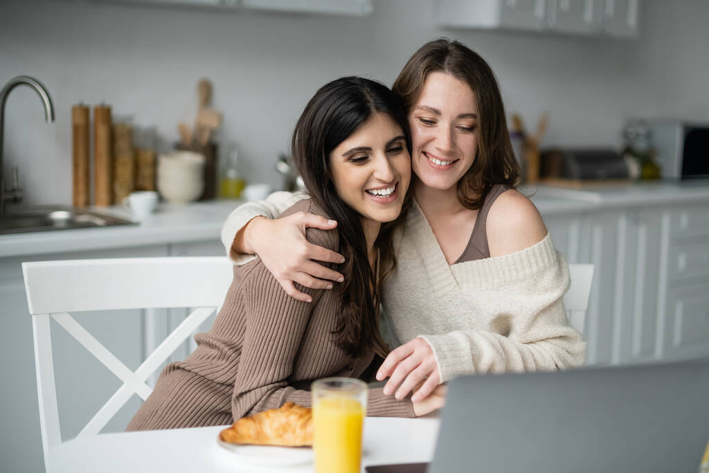Mujer sonriente en suéter abrazo compañero cerca de la computadora portátil y el desayuno en la cocina  - Foto, imagen