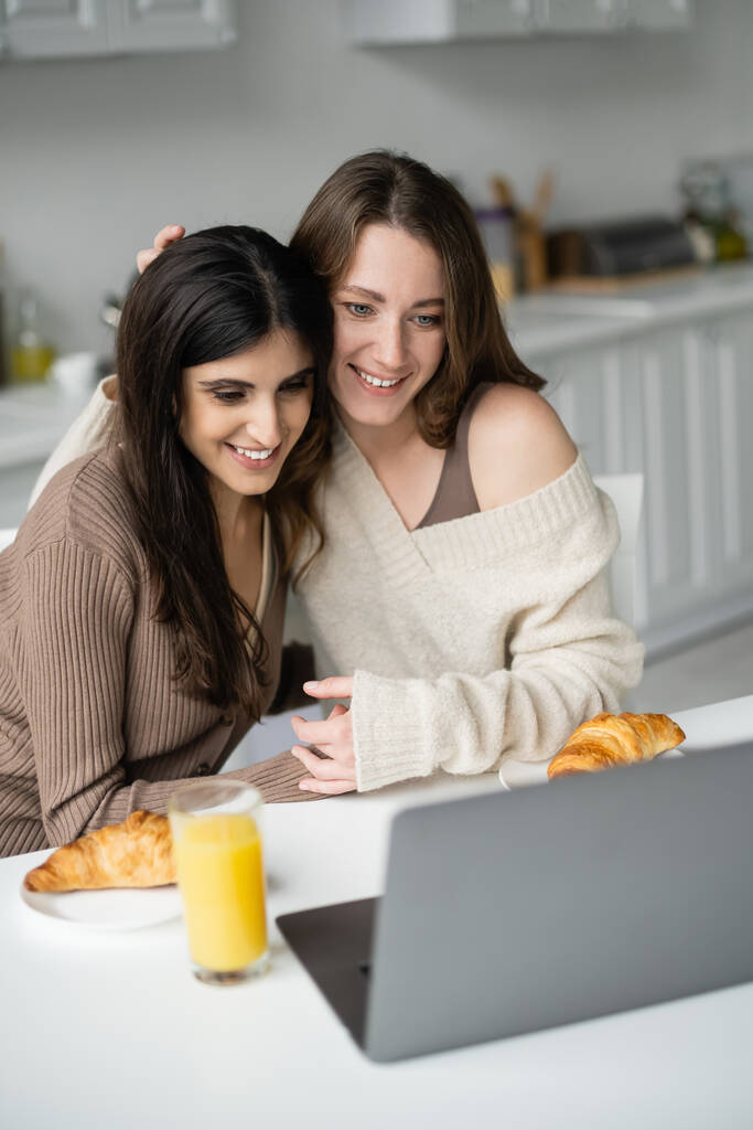 Gülümseyen kadın süveter giymiş, laptopun yanında sarılan eş ve mutfakta kahvaltı yapan.  - Fotoğraf, Görsel