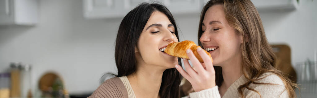 Positive lesbian woman feeding girlfriend with tasty croissant in kitchen, banner  - Photo, Image
