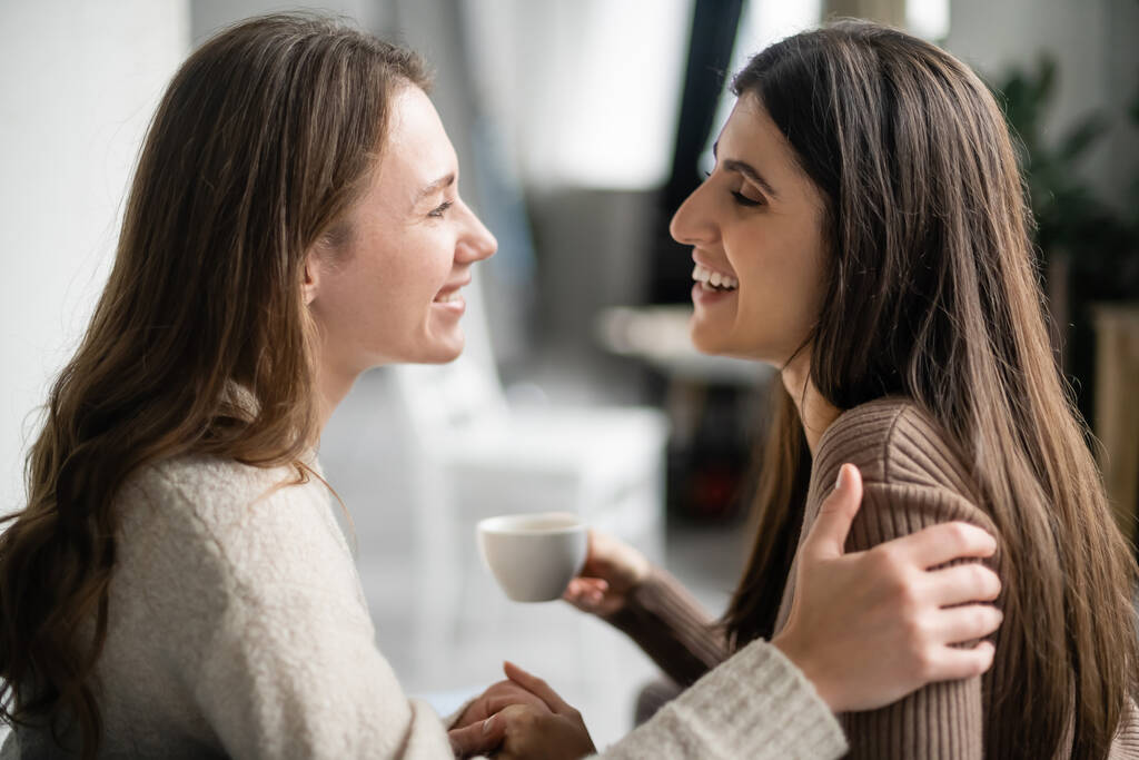 Вид сбоку лесбиянки в свитере, обнимающейся и разговаривающей с подругой за чашкой кофе дома  - Фото, изображение