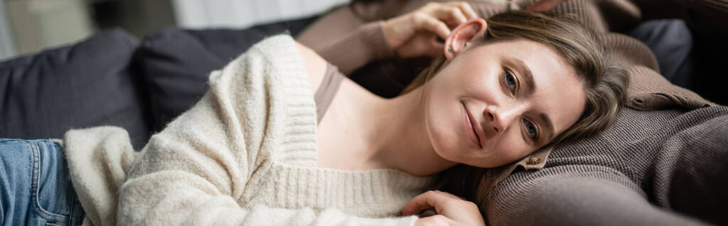 Femme souriante en pull regardant la caméra tout en étant couché près de la petite amie sur le canapé, bannière  - Photo, image