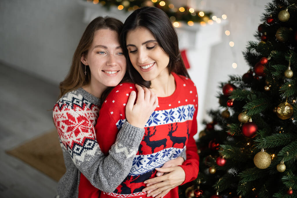 Glimlachende vrouw in warme trui aanraken partner in de buurt van kerstboom thuis  - Foto, afbeelding