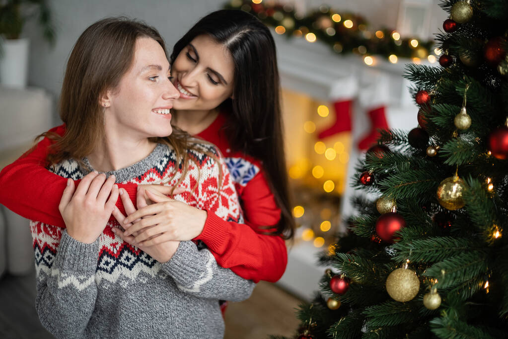 Young woman embracing girlfriend in sweater near decorated christmas tree  - Photo, Image