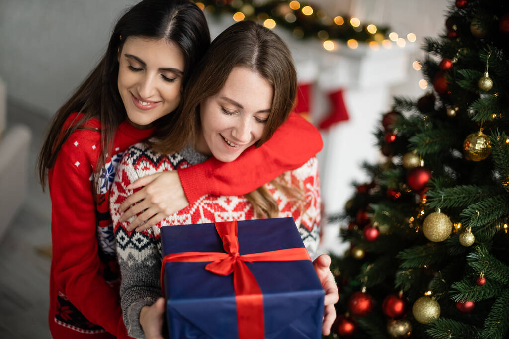 Positive Frau umarmt Freundin mit Geschenk neben Weihnachtsbaum zu Hause  - Foto, Bild