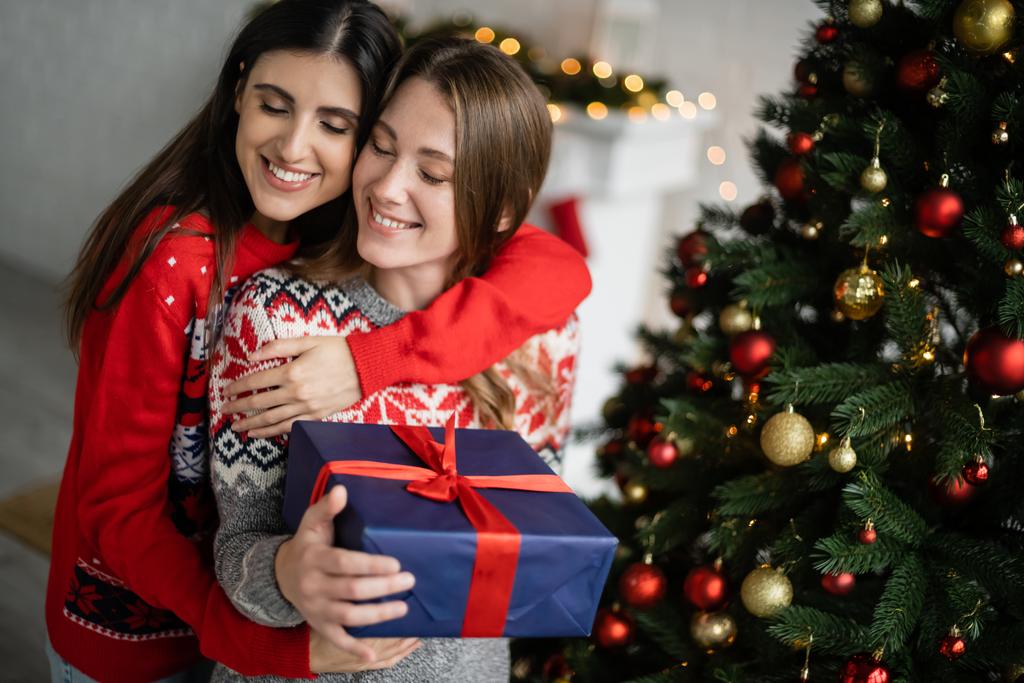 Mujer positiva abrazando novia con caja de regalo durante la celebración de Navidad en casa  - Foto, imagen