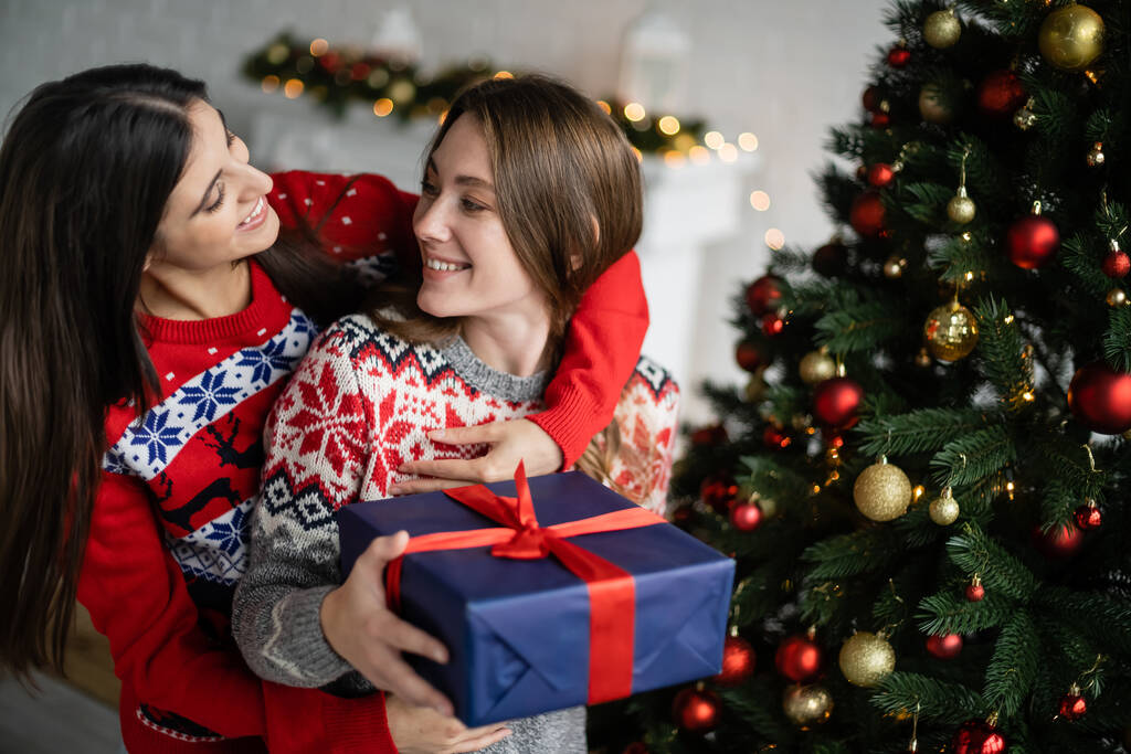 Positive Frau umarmt Freundin bei Neujahrsfeier im Wohnzimmer mit Geschenk  - Foto, Bild