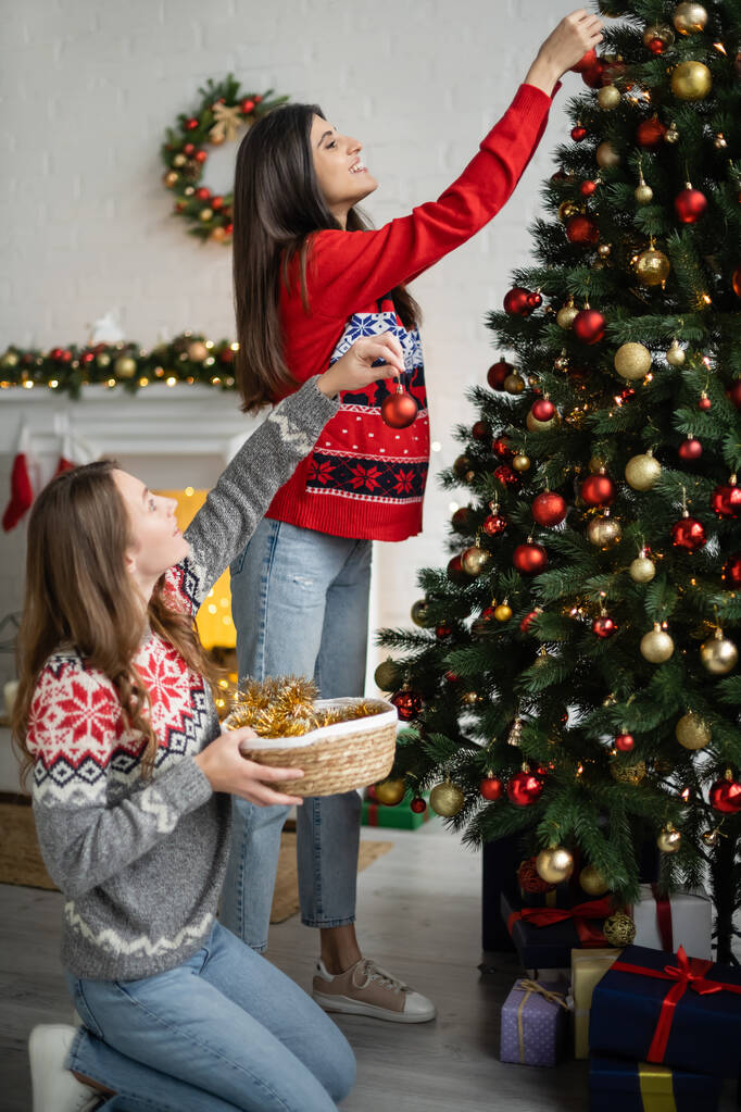Vista lateral de sonreír pareja del mismo sexo decorando árbol de Navidad en la sala de estar  - Foto, Imagen