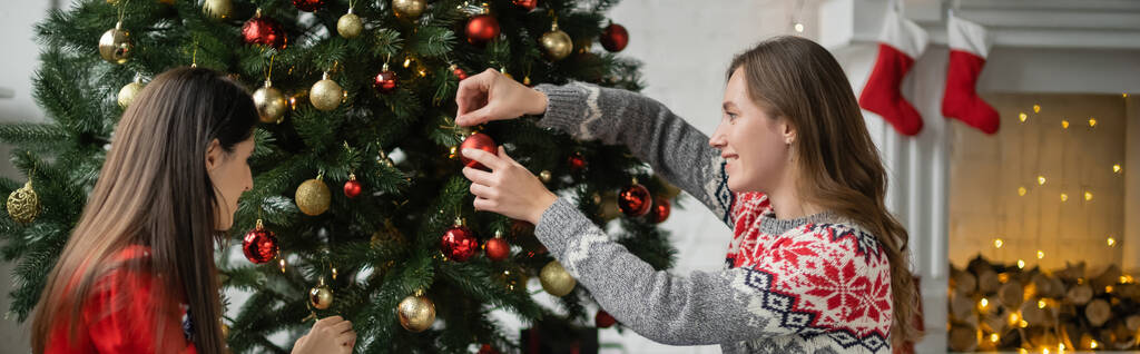 Vista laterale della coppia lesbica sorridente che decora l'albero di Natale con le palle a casa, banner  - Foto, immagini