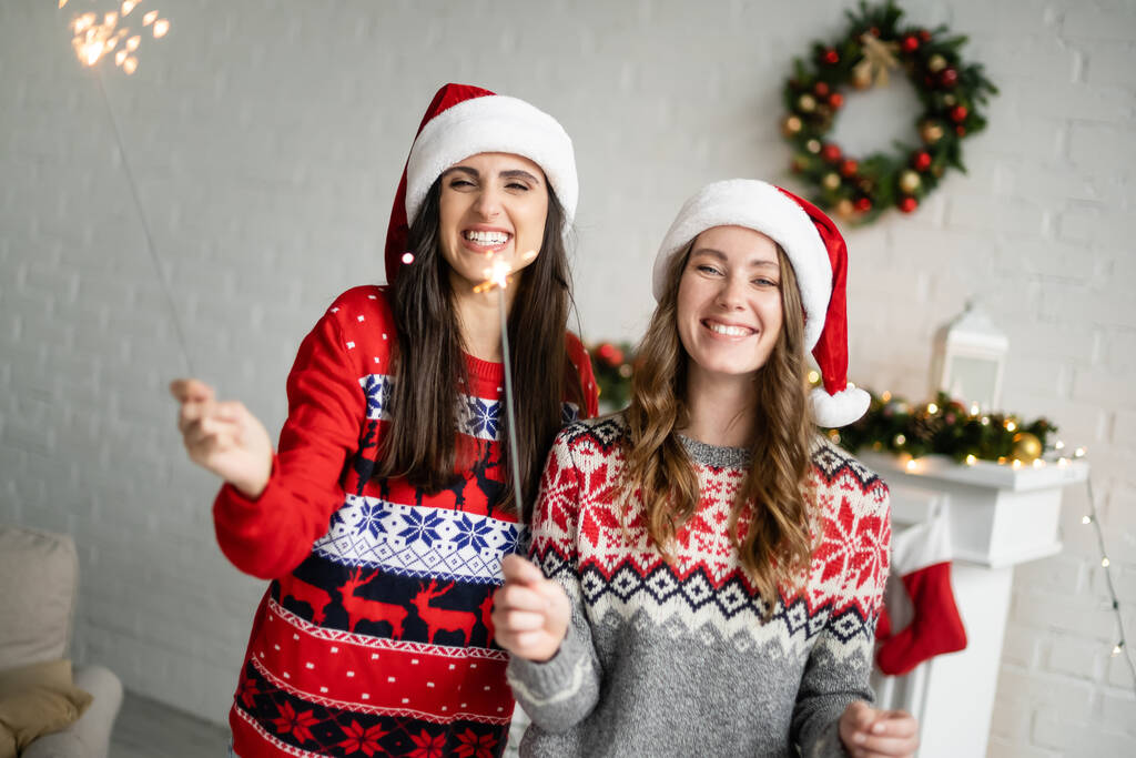 Felice coppia lesbica in cappelli di Babbo Natale in possesso di scintille in soggiorno  - Foto, immagini