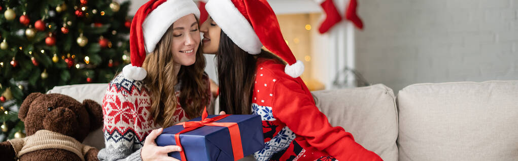 Mulher positiva em santa chapéu beijando namorada com presente durante a celebração do Natal em casa, banner  - Foto, Imagem