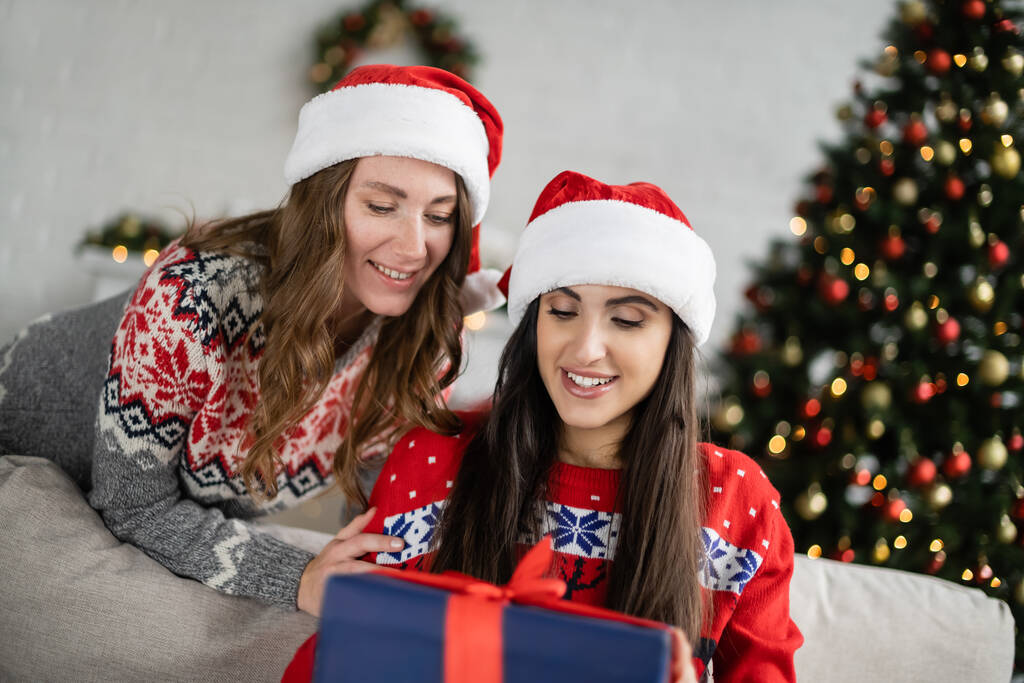 Positive lesbian couple in christmas gifts looking at blurred new year gift at home  - Photo, Image