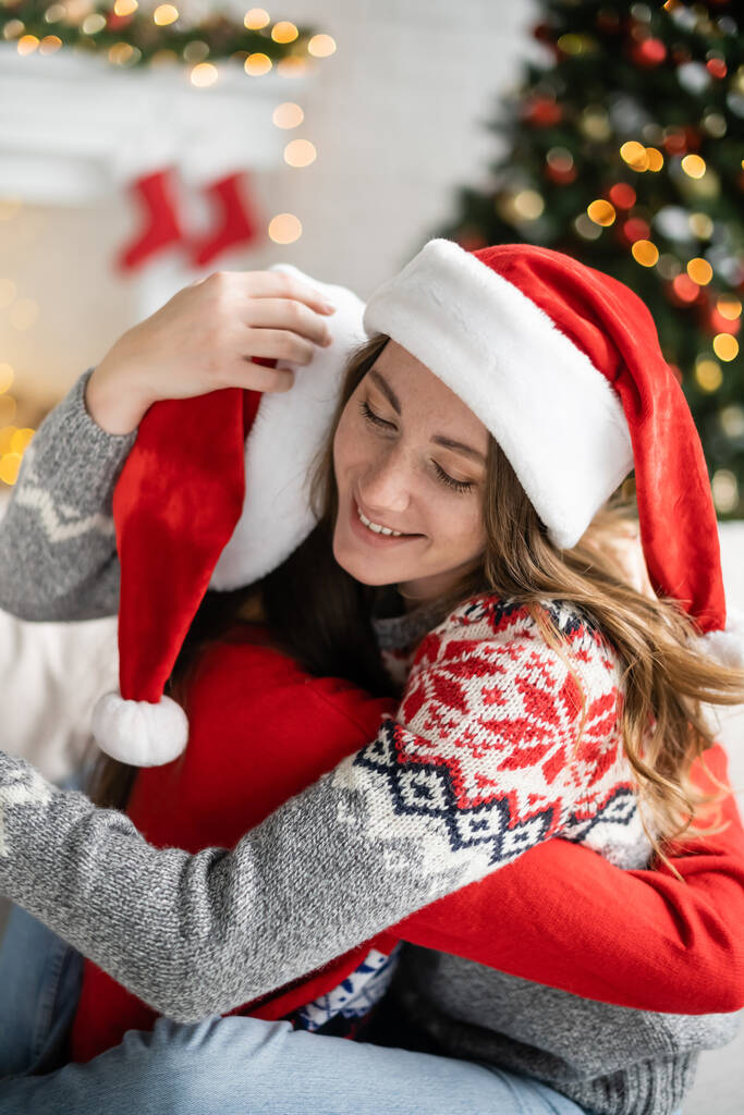 Donna sorridente in cappello da Babbo Natale che abbraccia la fidanzata durante la celebrazione del nuovo anno a casa  - Foto, immagini