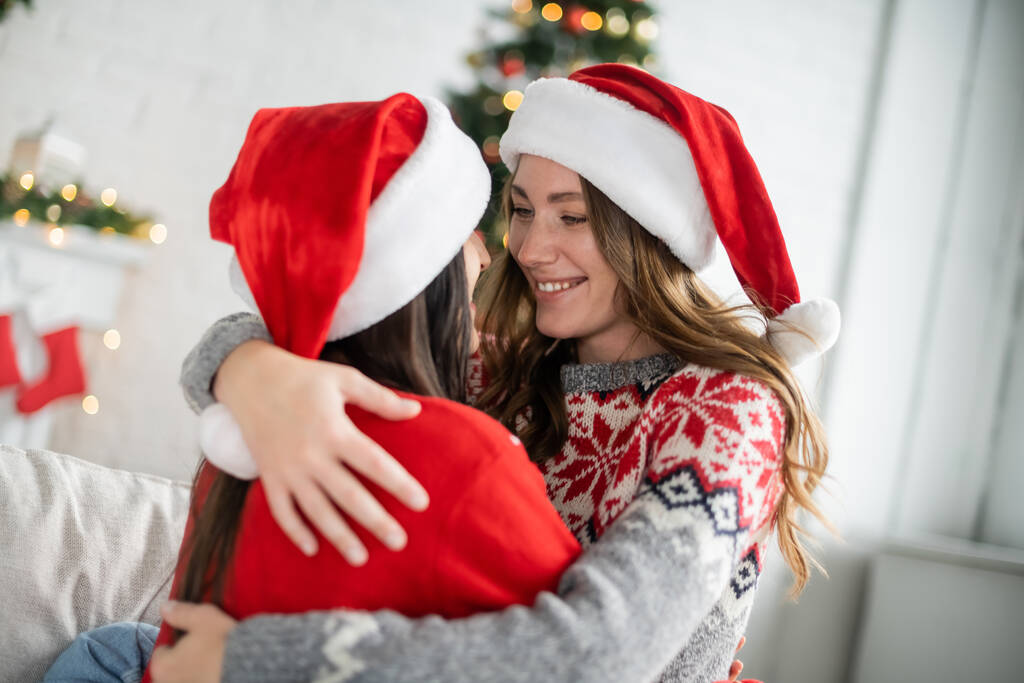 gelukkig lesbisch vrouw in santa hoed knuffelen vriendin op bank thuis  - Foto, afbeelding