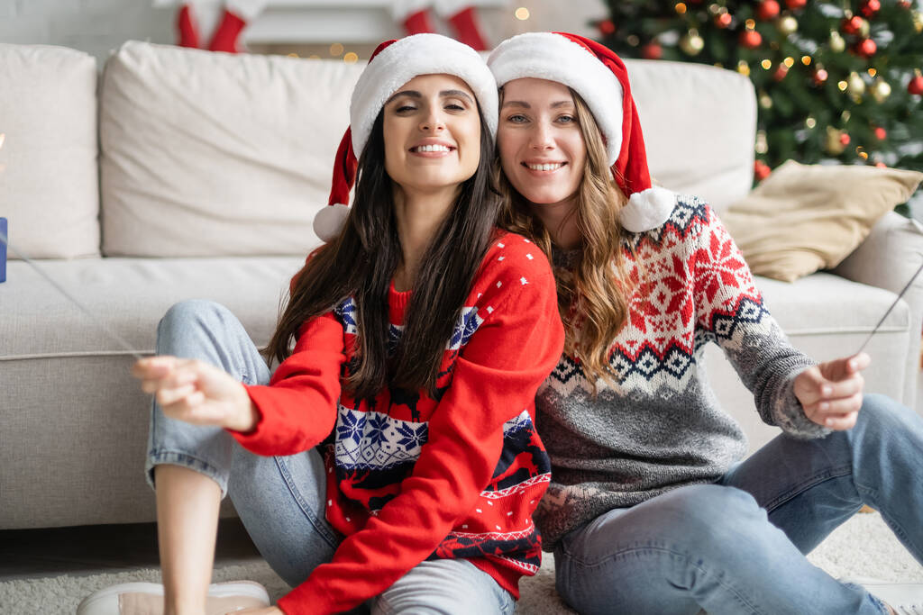 Positives lesbisches Paar mit Weihnachtsmann-Hüten und Wunderkerzen im Wohnzimmer  - Foto, Bild