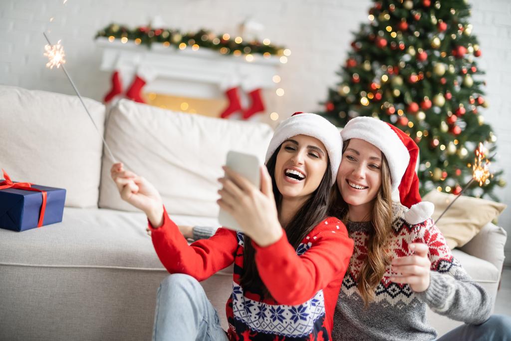 positif lesbienne couple dans santa chapeaux à l'aide de smartphone et tenant sparkers dans salon  - Photo, image