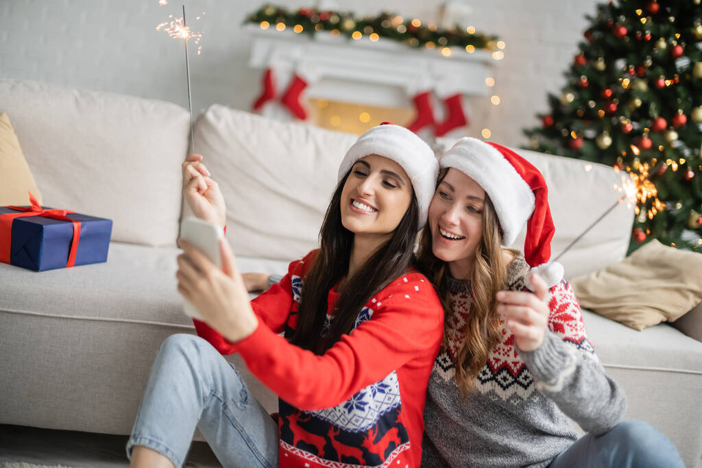 Positives lesbisches Paar nutzt Smartphone und hält Wunderkerzen bei Neujahrsfeier zu Hause  - Foto, Bild