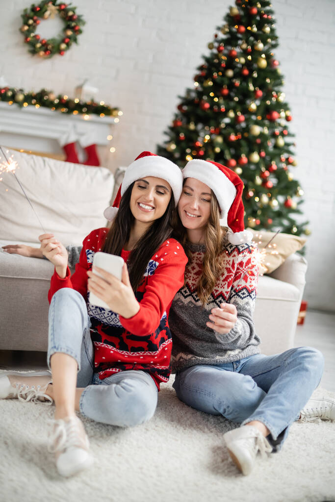 Giovane coppia lesbica in cappelli di Babbo Natale utilizzando il cellulare e tenendo scintille durante il Natale  - Foto, immagini