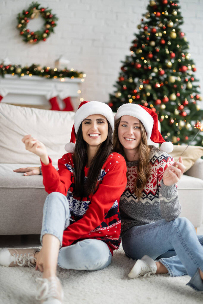 Fröhliches lesbisches Paar mit Weihnachtsmann-Hüten und Wunderkerzen bei der Silvesterfeier zu Hause  - Foto, Bild