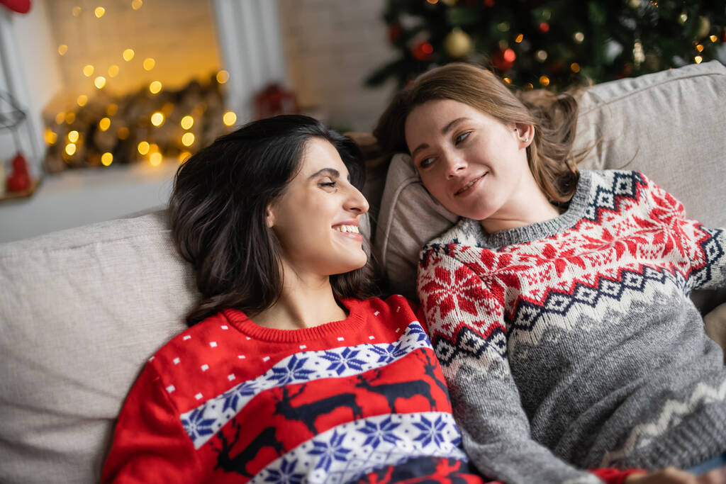 Позитивная лесбийская пара в новогодних свитерах отдыхает дома на диване  - Фото, изображение