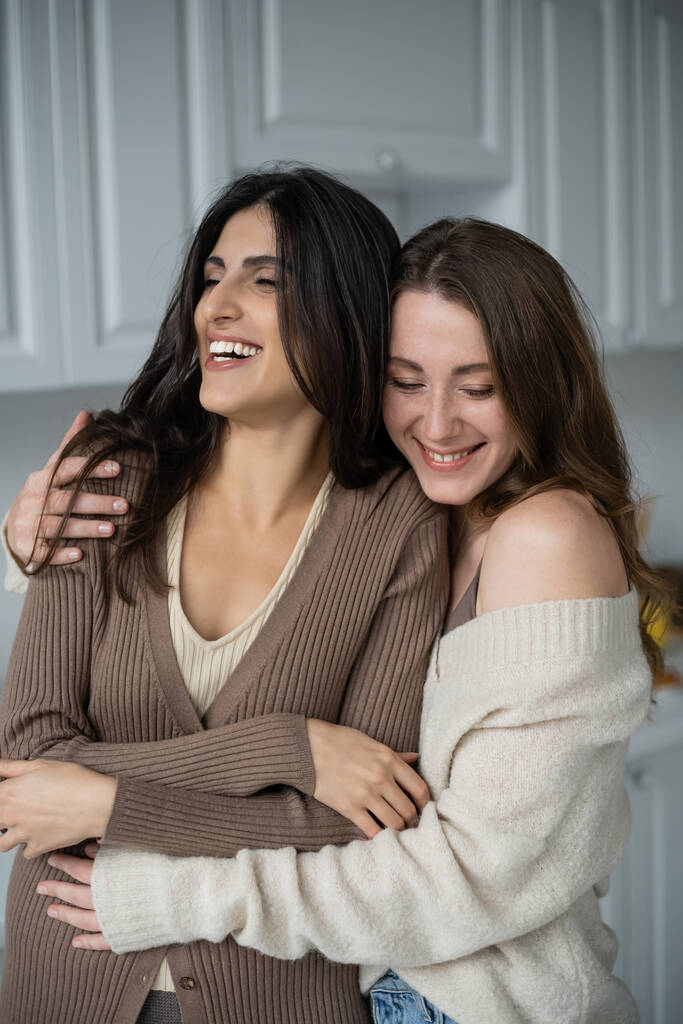 Fröhliche Frau im Pullover umarmt Freundin zu Hause  - Foto, Bild