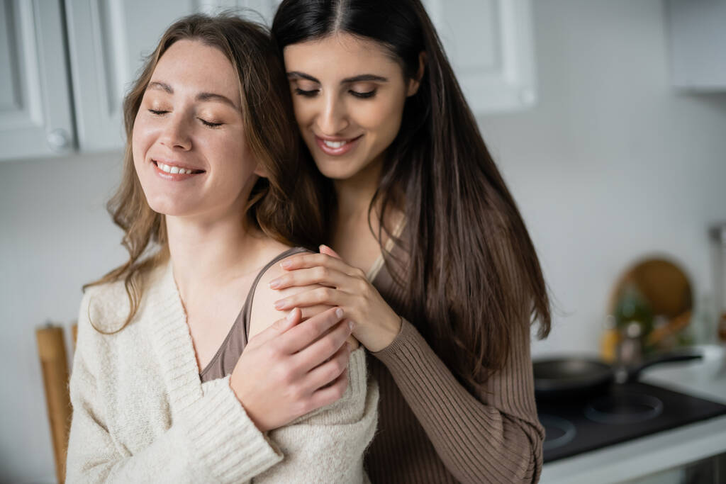 Pozitivní žena dotýká rameno přítelkyně ve svetru v kuchyni  - Fotografie, Obrázek
