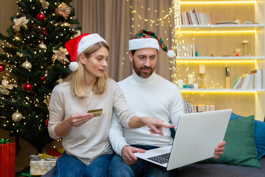 Feestelijke familie online winkelen. Een jong stel, een man en een vrouw, zitten thuis op de bank in de buurt van de kerstboom in rode hoeden, met een laptop en een creditcard. - Foto, afbeelding