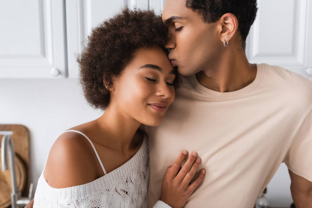 giovane africano americano uomo baci sorridente fidanzata in piedi con gli occhi chiusi in cucina - Foto, immagini
