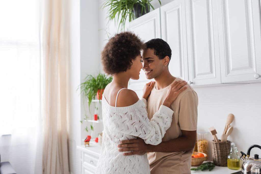 frisé afro-américain femme en blanc ajouré pull souriant près heureux petit ami dans la cuisine - Photo, image