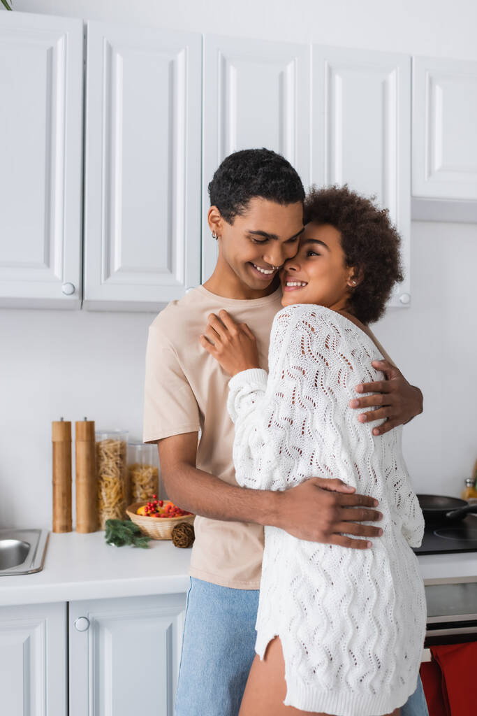μελαχρινή αφροαμερικάνικη γυναίκα σε λευκό πλεκτό πουλόβερ αγκαλιάζει με το νεαρό φίλο της στην κουζίνα - Φωτογραφία, εικόνα