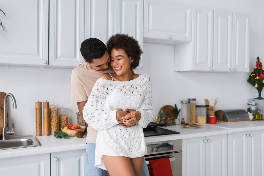 Afrikaans amerikaanse man knuffelen vrolijke vriendin in wit gebreide trui in keuken - Foto, afbeelding