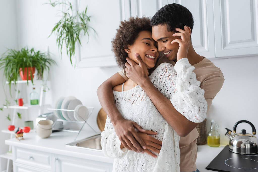 allegra donna afroamericana in maglione bianco lavorato a maglia che abbraccia con il giovane fidanzato in cucina - Foto, immagini