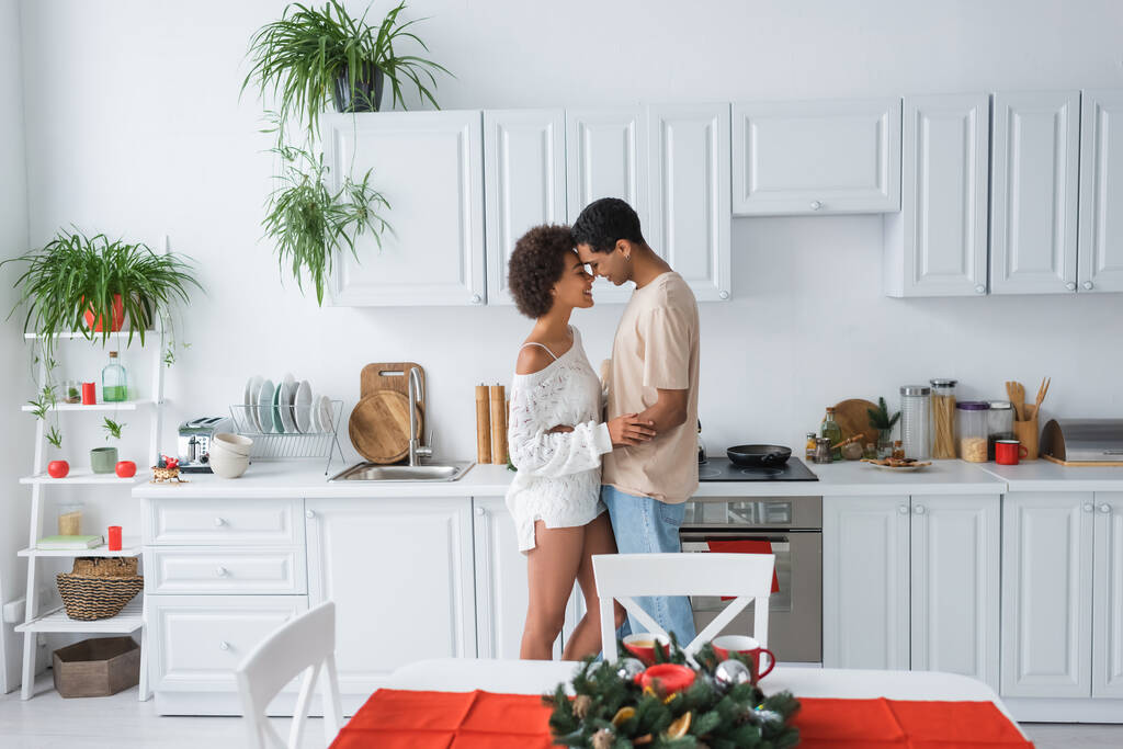 πλαϊνή όψη του σέξι Αφροαμερικανού ζευγαριού αγκαλιάζει στη σύγχρονη κουζίνα με λευκά έπιπλα - Φωτογραφία, εικόνα