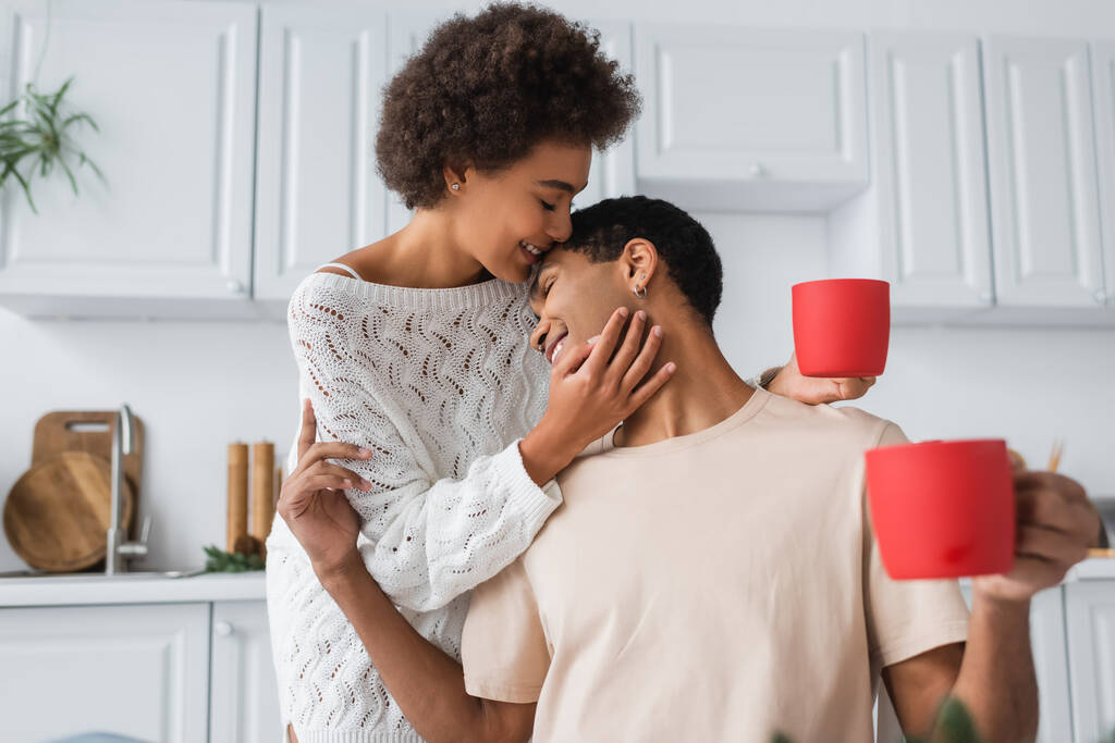 potěšený africký americký pár s červenými poháry objímání v kuchyni - Fotografie, Obrázek