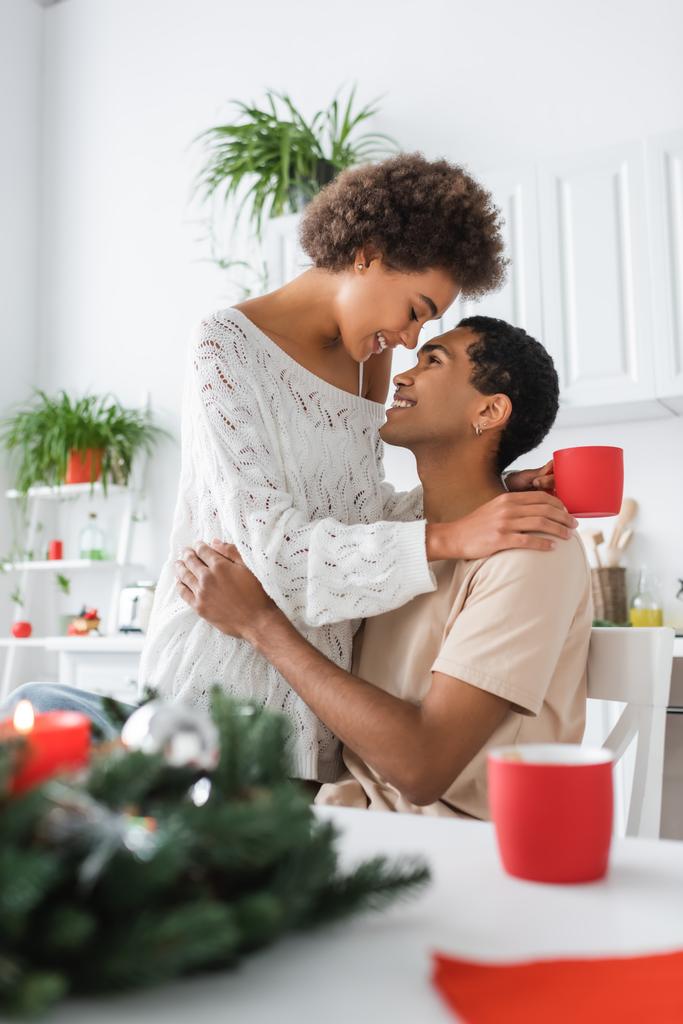 smiling african american woman with red cup hugging young brunette boyfriend in kitchen - Photo, Image