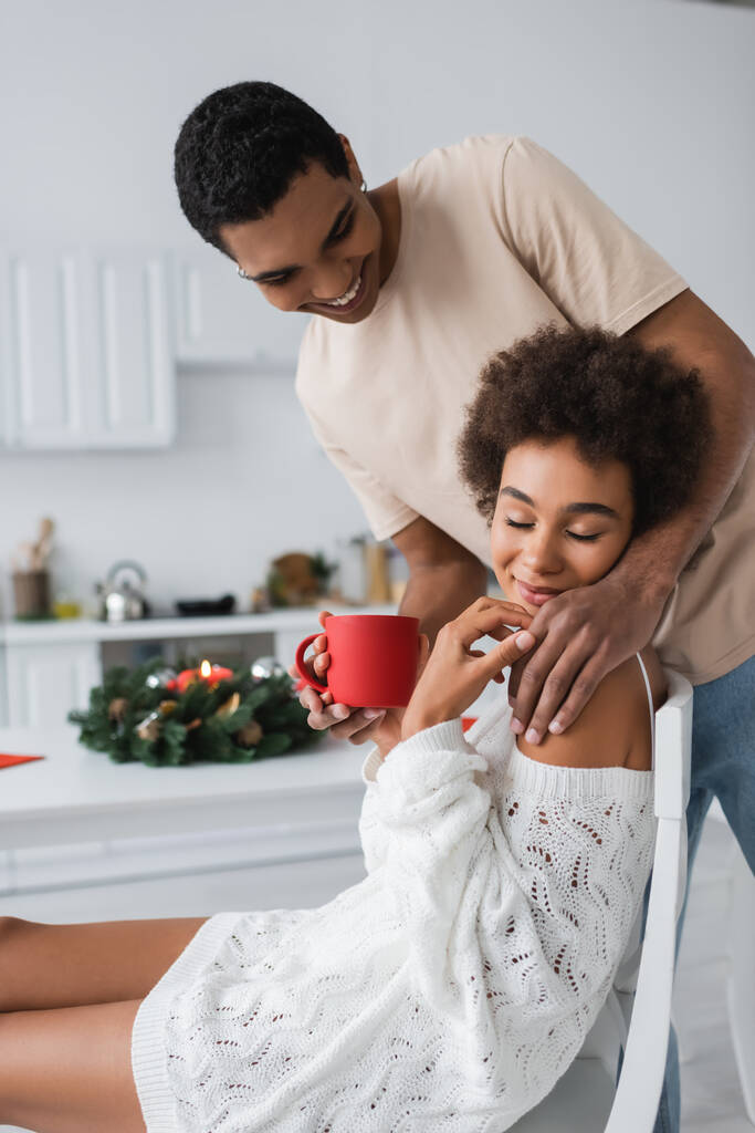 smiling african american man with red cup touching seductive girlfriend smiling with closed eyes in kitchen - Photo, Image