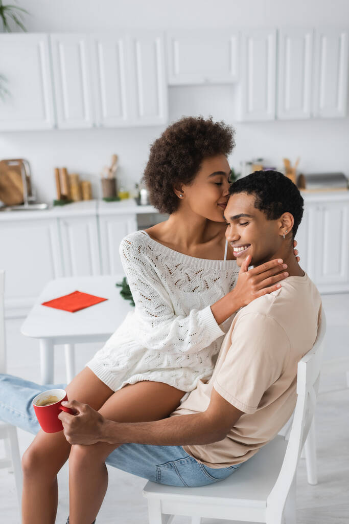 sensual african american woman hugging boyfriend sitting in kitchen with red cup - Photo, Image