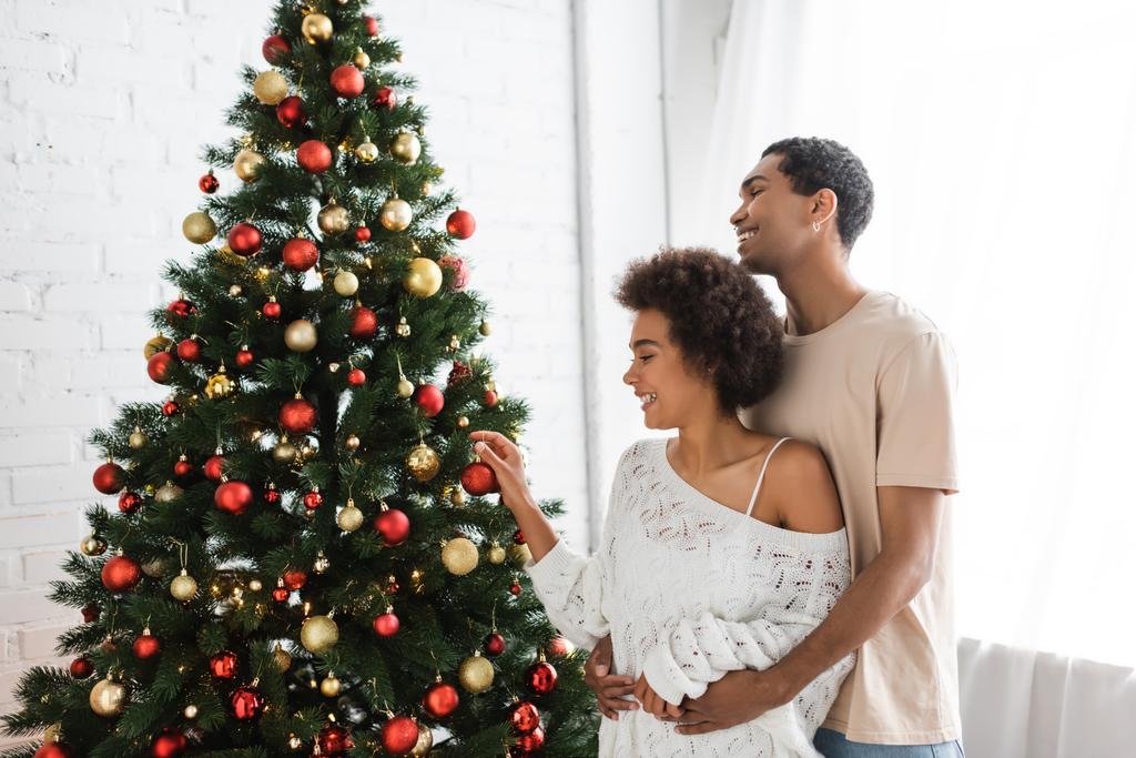 homme afro-américain étreignant petite amie en chandail ajouré blanc décorant sapin avec des boules de Noël - Photo, image