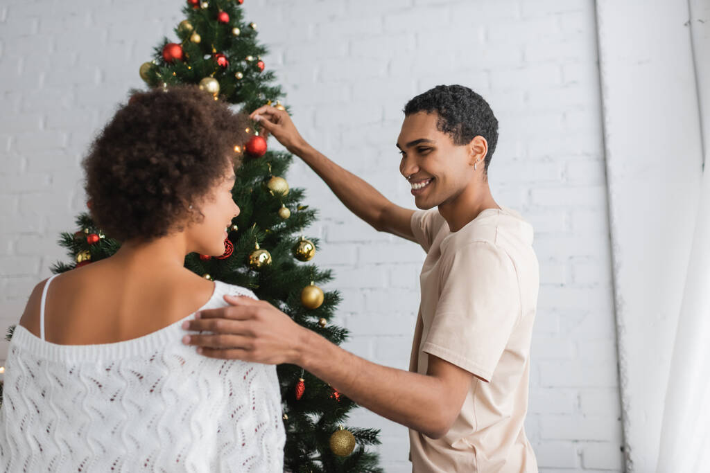 Neşeli Afrikalı Amerikalı adam beyaz önlüklü kazak ve Noel ağacı süsleme kız arkadaşına dokunuyor. - Fotoğraf, Görsel