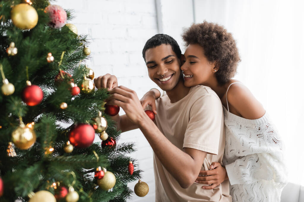 sexy africký americký žena objímající šťastný přítel zdobení borovice s vánoční koule - Fotografie, Obrázek
