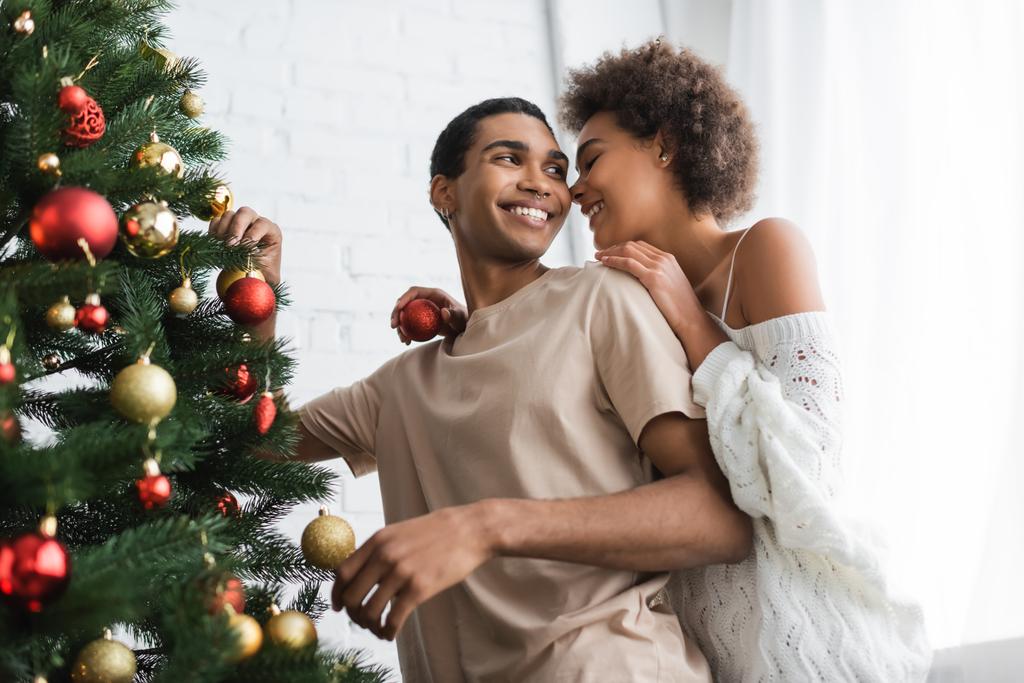 若いですアフリカ系アメリカ人女性は笑顔のボーイフレンドの近くにクリスマスボールを保持松の木を飾る - 写真・画像
