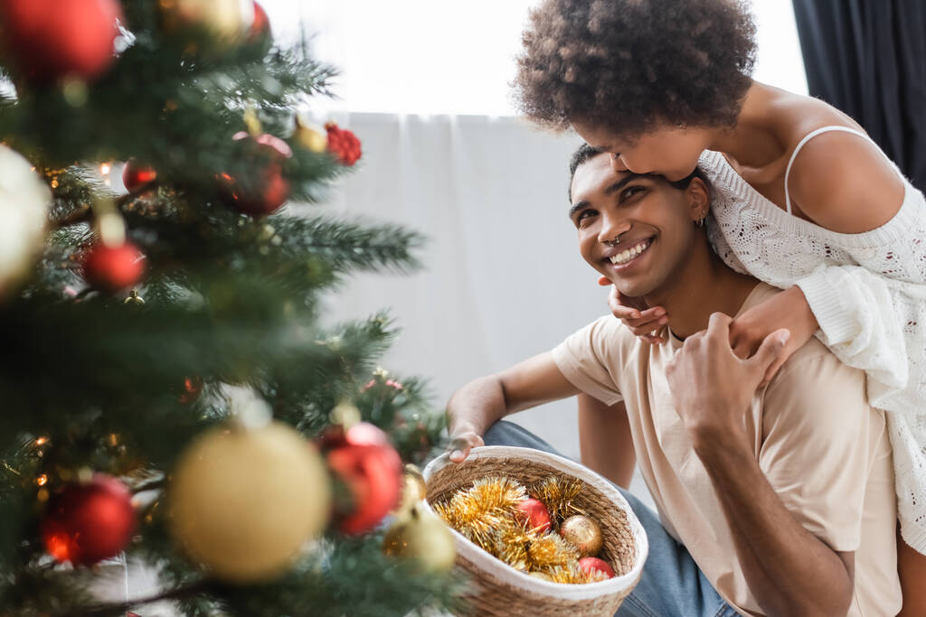ricci donna africana americana abbracciando allegro fidanzato tenendo cesto di vimini con le bagattelle vicino albero di Natale - Foto, immagini