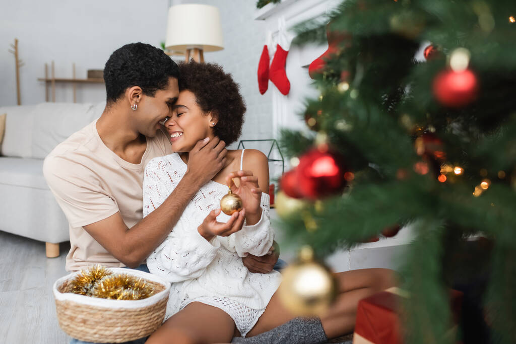 giovane uomo afro-americano che abbraccia donna felice con bauble vicino all'albero di Natale offuscato - Foto, immagini