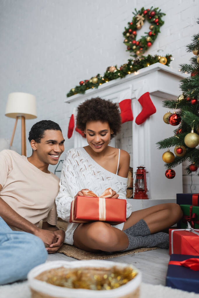 χαρούμενη και σέξι Αφροαμερικανή που κάθεται στο πάτωμα με χριστουγεννιάτικο δώρο κοντά στο τζάκι και χαμογελαστό αγόρι  - Φωτογραφία, εικόνα