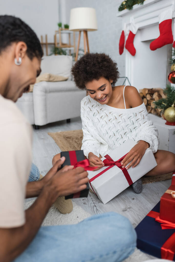 feliz africano americano pareja apertura navidad regalos mientras sentado en piso en casa - Foto, Imagen